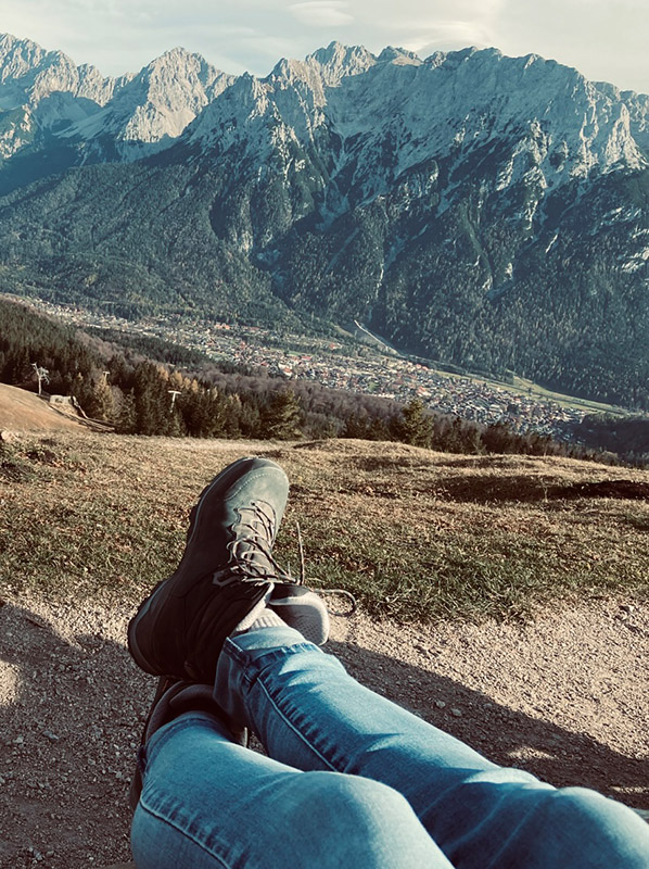 Foto mit einer Aussicht auf Berge und im Vordergrund Beine mit Wanderschuhen